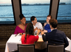Five people sit around a dining table on a boat, smiling and raising glasses in a toast. A large window shows a view of the ocean in the background.