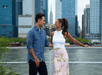 Two people stand on a waterfront, smiling at each other. The city skyline, including the Empire State Building, is visible in the background.