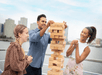 Three people play a game of giant Jenga on a boat deck, with a city skyline in the background.