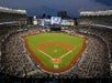 Aerial view looking down on Yankee Stadium with thousands of fans filling the stands and players on the field at dusk.
