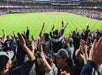 Yankees fans in the stands cheering and jumping with their arms in the air with the bright green field in front of them.