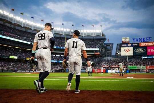 Two New York Yankees baseball players walk side by side near the field during a game at Yankee Stadium, with fans in the stands and the sky above.