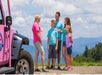 A tour guide in a pink shirt is speaking to a family of four near a pink off-road vehicle, with mountains visible in the background.