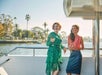 Two women smiling and holding drinks standing on the desk of the Newport Beach Brunch Cruise with the sun shining behind them.