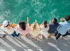 A line of people holding drinks looking over the railing of the boat into the blue green water on a sunny day on the Newport Beach Brunch Cruise.