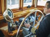 A ship captain in a suit turning a giant gold steering wheel in the bridge of the Newport Beach Brunch Cruise.