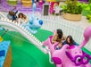 Families ride a colorful amusement park ride themed with pink and blue dog characters, surrounded by white fencing and vibrant decor.