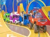 Children and adults ride in colorful, toy-like cars on a track indoors, surrounded by bright blue and yellow decorations.