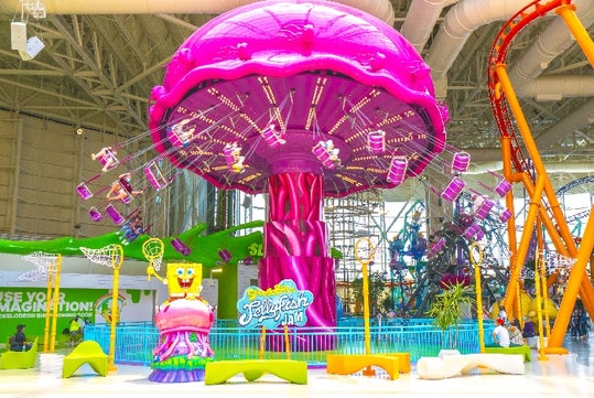 A bright pink jellyfish-themed swing ride at an indoor amusement park, with SpongeBob SquarePants decor and colorful seating in the foreground.