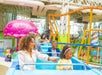 A family enjoys a ride on a blue roller coaster at an indoor amusement park. The background features colorful structures and a pink rocket-shaped decor.