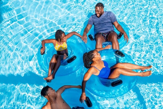 Four people relax on blue inflatable rings in a pool, smiling and chatting under the sun.