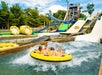 Four people ride a yellow circular raft down a water slide at an amusement park. In the background, other slides are visible with spectators watching.
