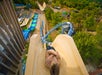 Person sliding down a tall water slide at an amusement park, surrounded by green trees and other attractions visible in the background.