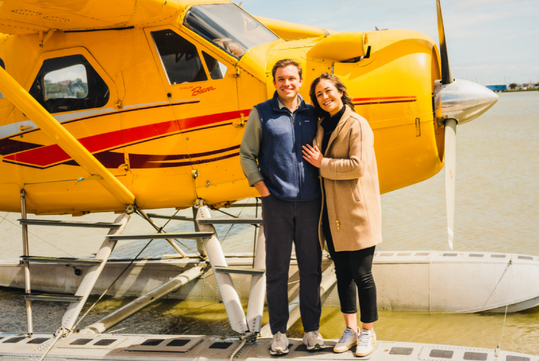 A smiling couple stands in front of a yellow floatplane docked on the water.