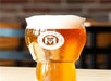 A glass of foamy beer with a logo on a wooden table, against a blurred background.