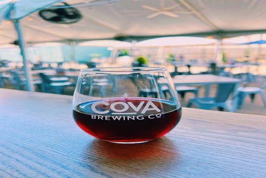 Tented outdoor seating area with a glass of dark beverage on a wooden table, marked with "COVA Brewing Co".