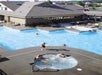 Aerial view of people swimming in a large outdoor pool and a smaller circular hot tub by a building.