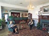 Two women sit in armchairs in a hotel lobby with a patterned carpet, fireplace, chandelier, and reception desk in the background.