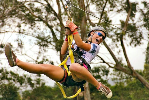 A person wearing a helmet and sunglasses is zip-lining outdoors, surrounded by trees, with an expression of excitement.