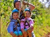 An adult and two children wearing helmets are harnessed together while zip-lining through a forested area.