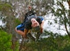 A person ziplines upside down with a big smile in a forested area.