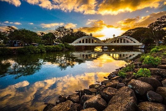 A bridge with arched supports spans across a river, reflecting the vibrant sunset sky, surrounded by greenery and rocks along the riverbank.