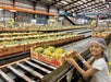A child in a hairnet smiles next to a conveyor belt filled with boxes of pineapples in a large warehouse.