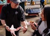 A smiling vendor hands food to a woman, who is holding a wrapped sandwich and laughing at an outdoor food stand.