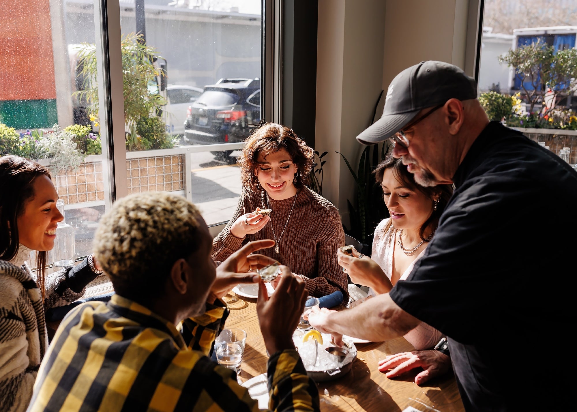 A group of four people sit at a table by a window while a server hands them food, with sunlight streaming into the room.