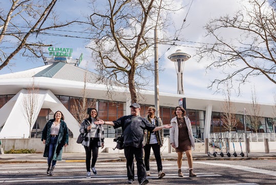 Five people cross a street in front of Seattle’s Climate Pledge Arena, with the Space Needle visible in the background on a partly cloudy day.