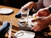 A person serves a metal dish of oysters on ice with lemon wedges at a wooden table set with plates, napkins, and glasses of water.