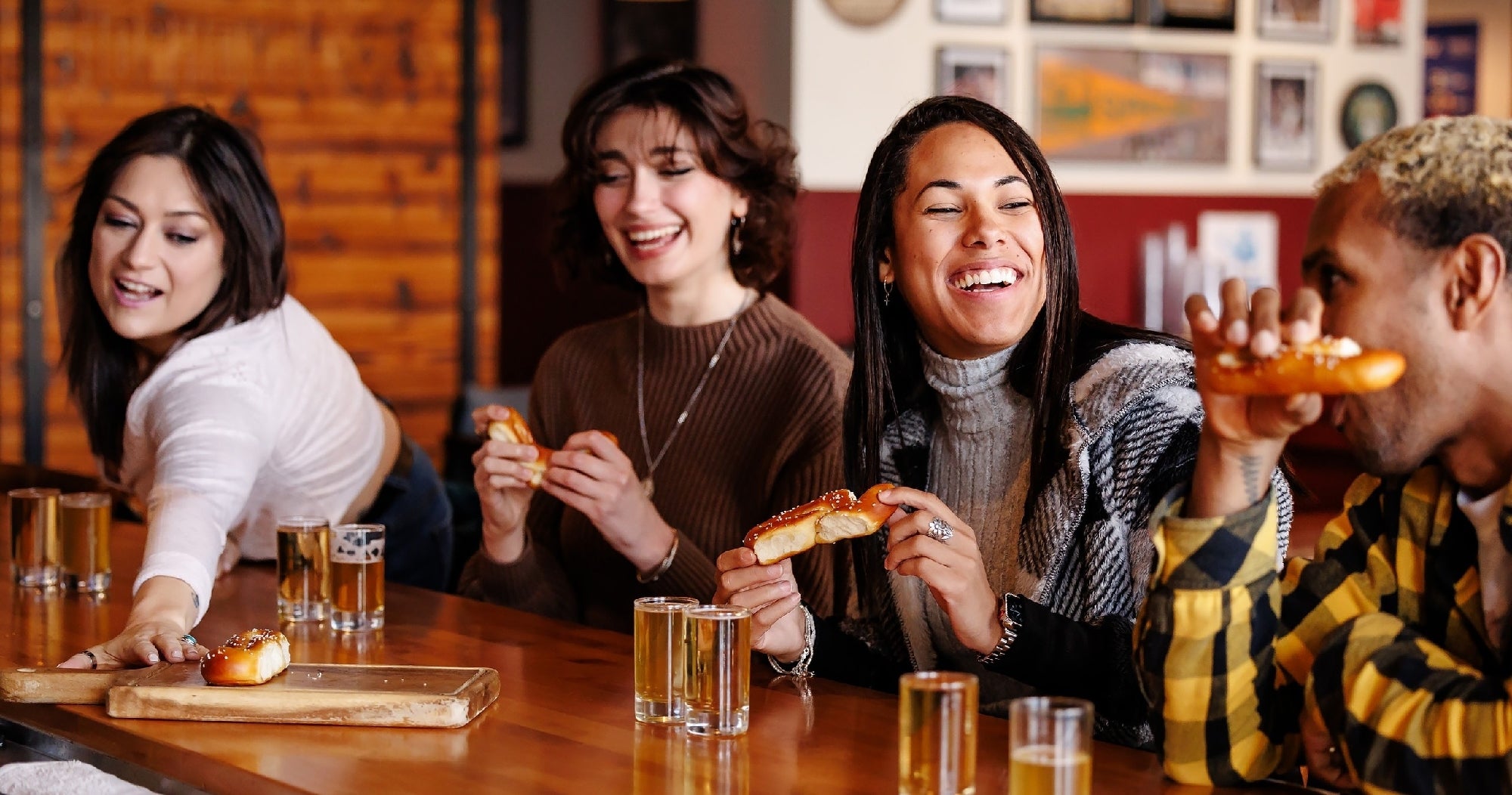 Four people sit at a bar enjoying soft pretzels and drinks, smiling and engaging with each other in a casual, social setting.