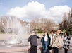 A group of five people stands and talks near a large outdoor fountain with a rainbow visible. Trees and radio towers are in the background under a partly cloudy sky.