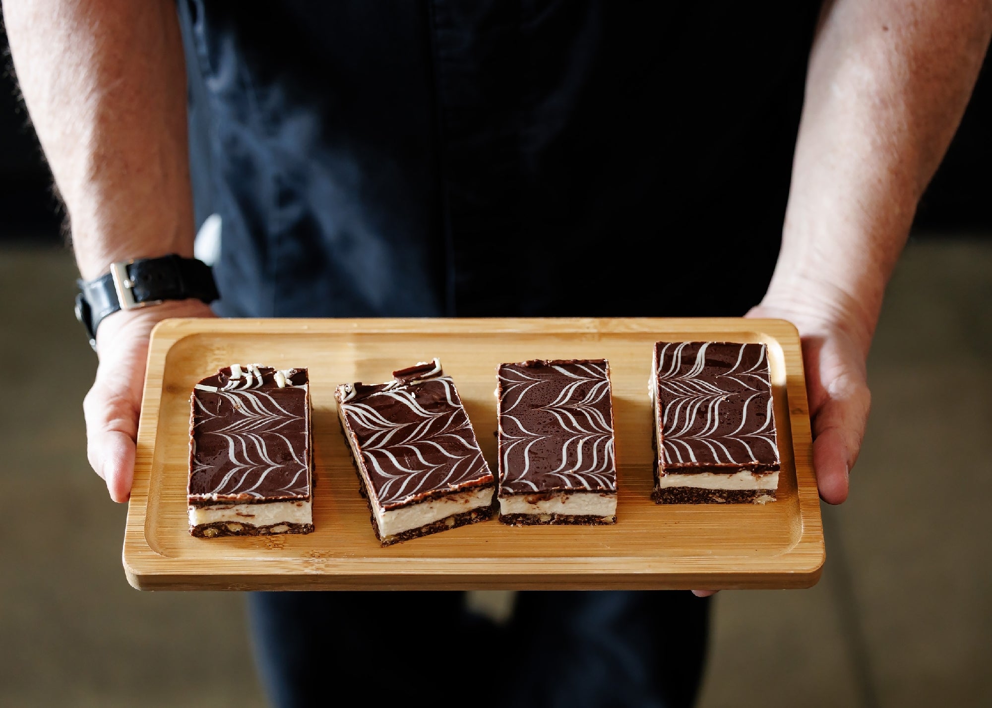 Person holding a wooden tray with four rectangular chocolate layered dessert bars, each topped with a marbled pattern.
