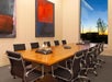 A modern conference room with a large wooden table, surrounded by black chairs. Abstract art hangs on the walls, and a window reveals a sunset view outside.
