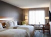 Hotel room with two double beds, a desk with chair, a flat-screen TV, and a large window with beige curtains. Room features modern decor with neutral tones.