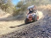 Two people drive an off-road ATV around a dusty dirt trail bend, with dry grass and trees in the background.