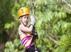 A child wearing a helmet and gloves rides a zipline through a green forested area.