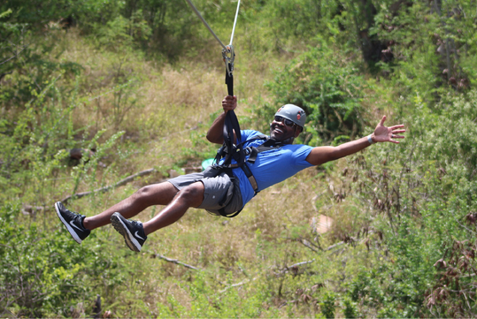 A man wearing a helmet and harness is ziplining outdoors over greenery, with one arm extended and smiling.