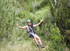 A person wearing a helmet and harness is ziplining through a lush green forest with arms outstretched and smiling.