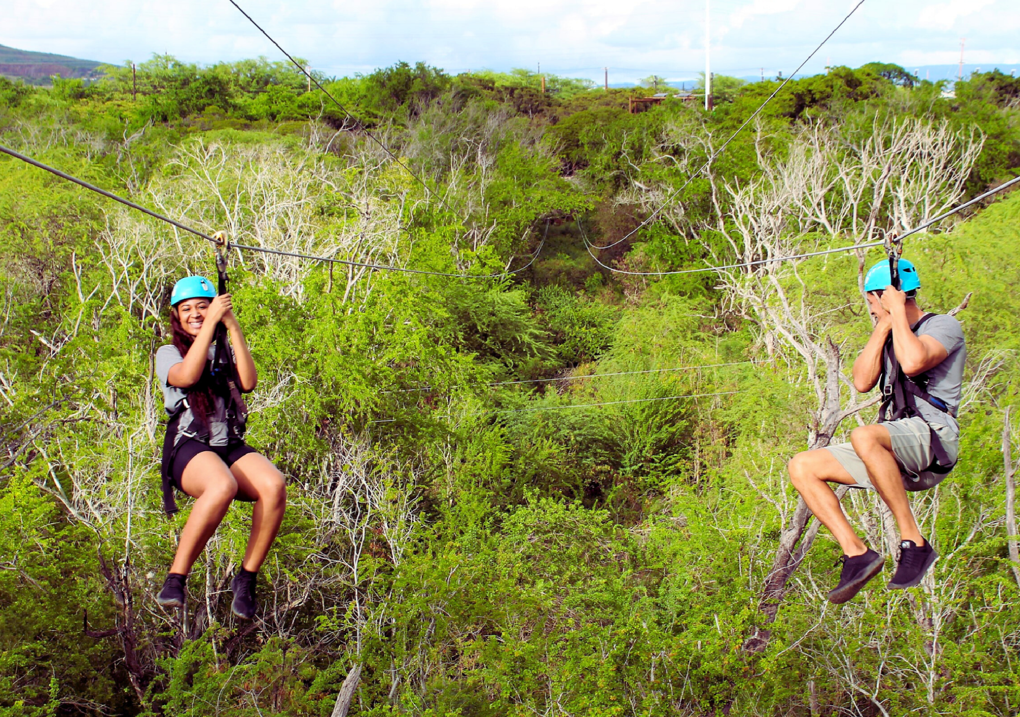 Two people wearing helmets ride parallel zip lines over a green forested area, one smiling at the camera and the other covering their face.