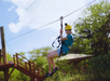 Person wearing a yellow helmet and safety gear rides a zip line outdoors, smiling, with trees and a wooden structure in the background.