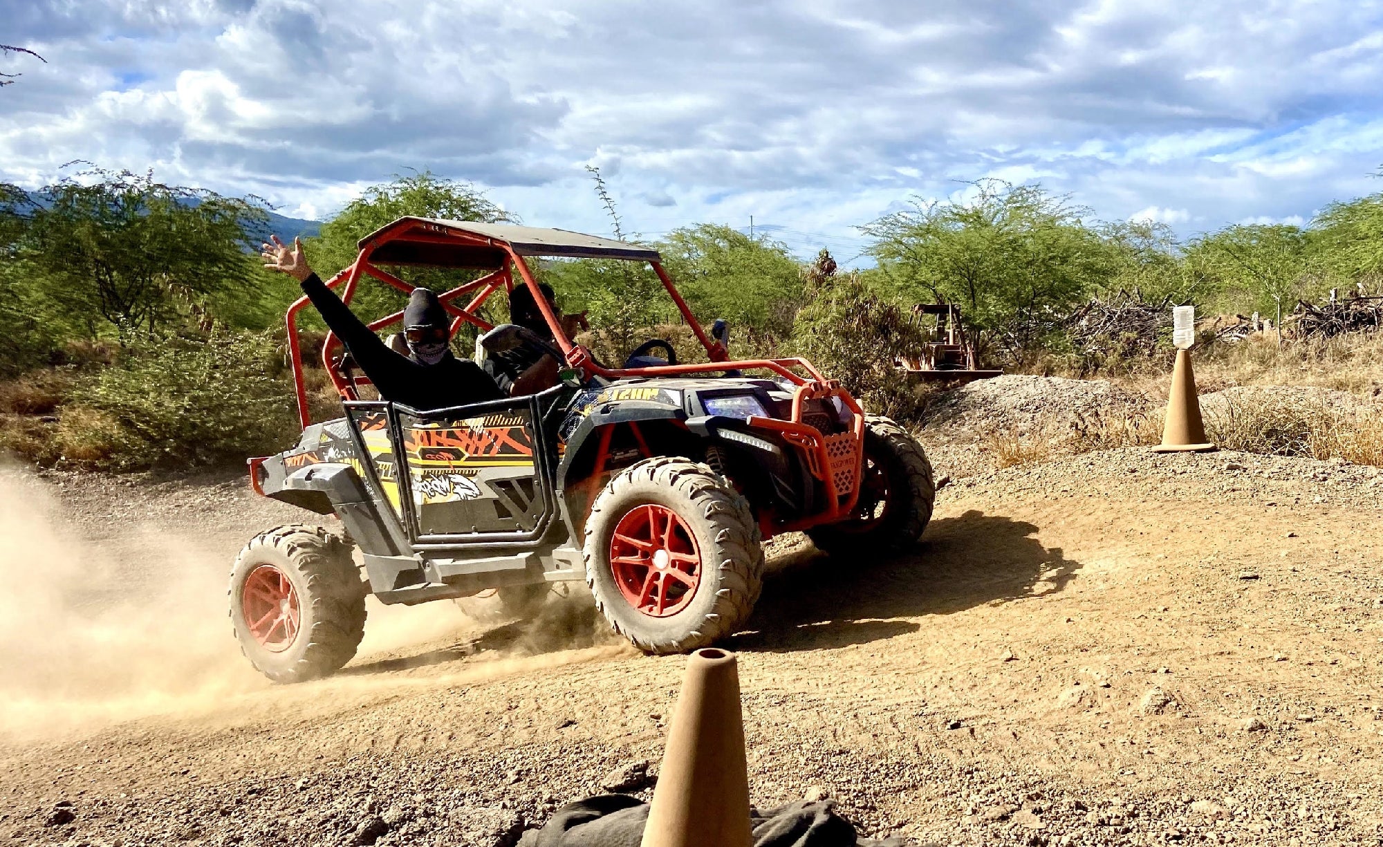 Two people drive an off-road vehicle on a dusty dirt trail surrounded by shrubs and trees, with orange cones marking the path.