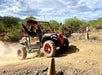 Two people drive an off-road vehicle on a dusty dirt trail surrounded by shrubs and trees, with orange cones marking the path.