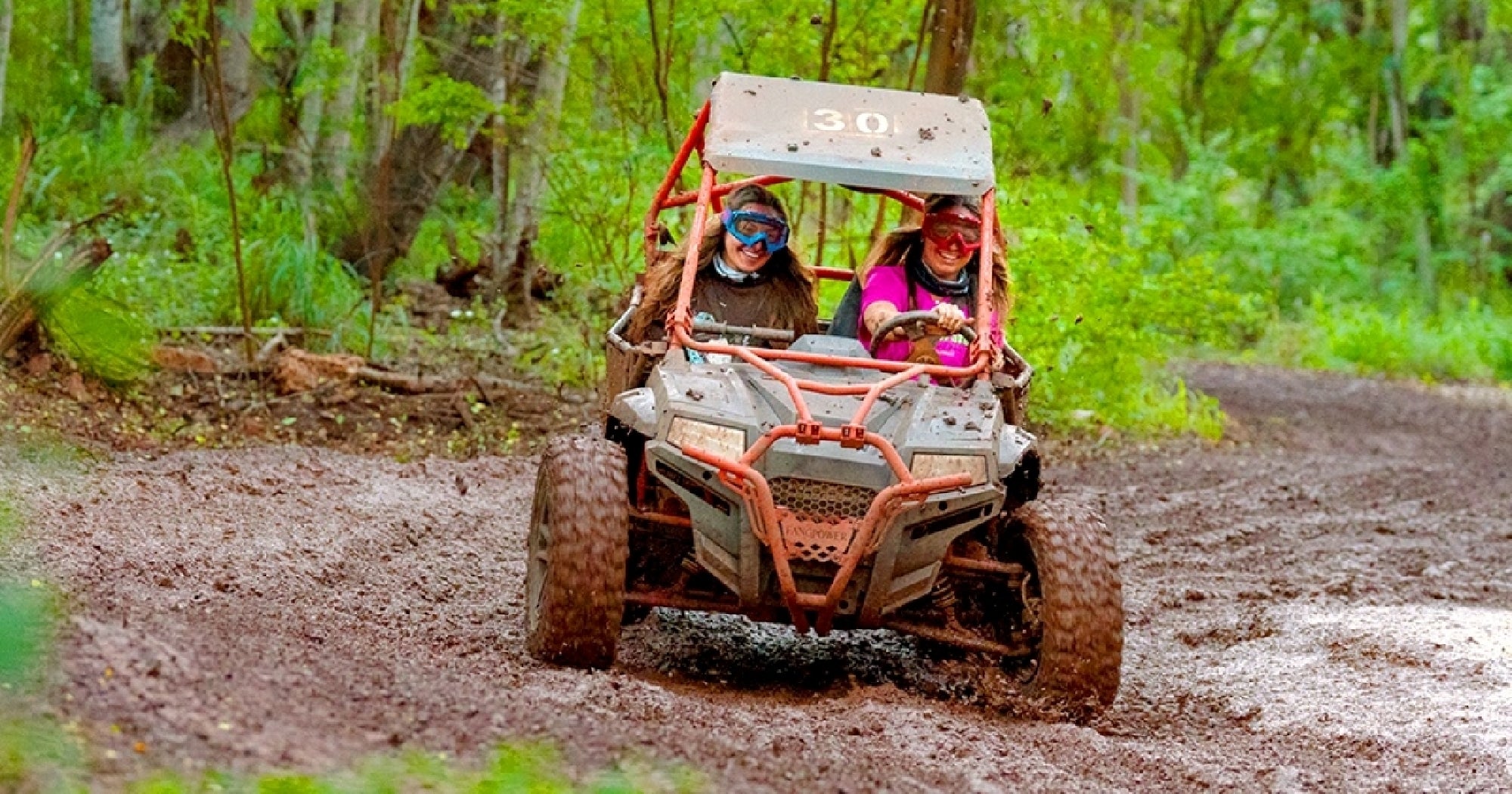 Two people wearing helmets and goggles drive an off-road buggy through a muddy trail surrounded by green trees.