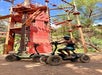 Person in helmet and sunglasses sits on an off-road electric scooter near a climbing tower and rope course, with another scooter beside them, outdoors on a sunny day.