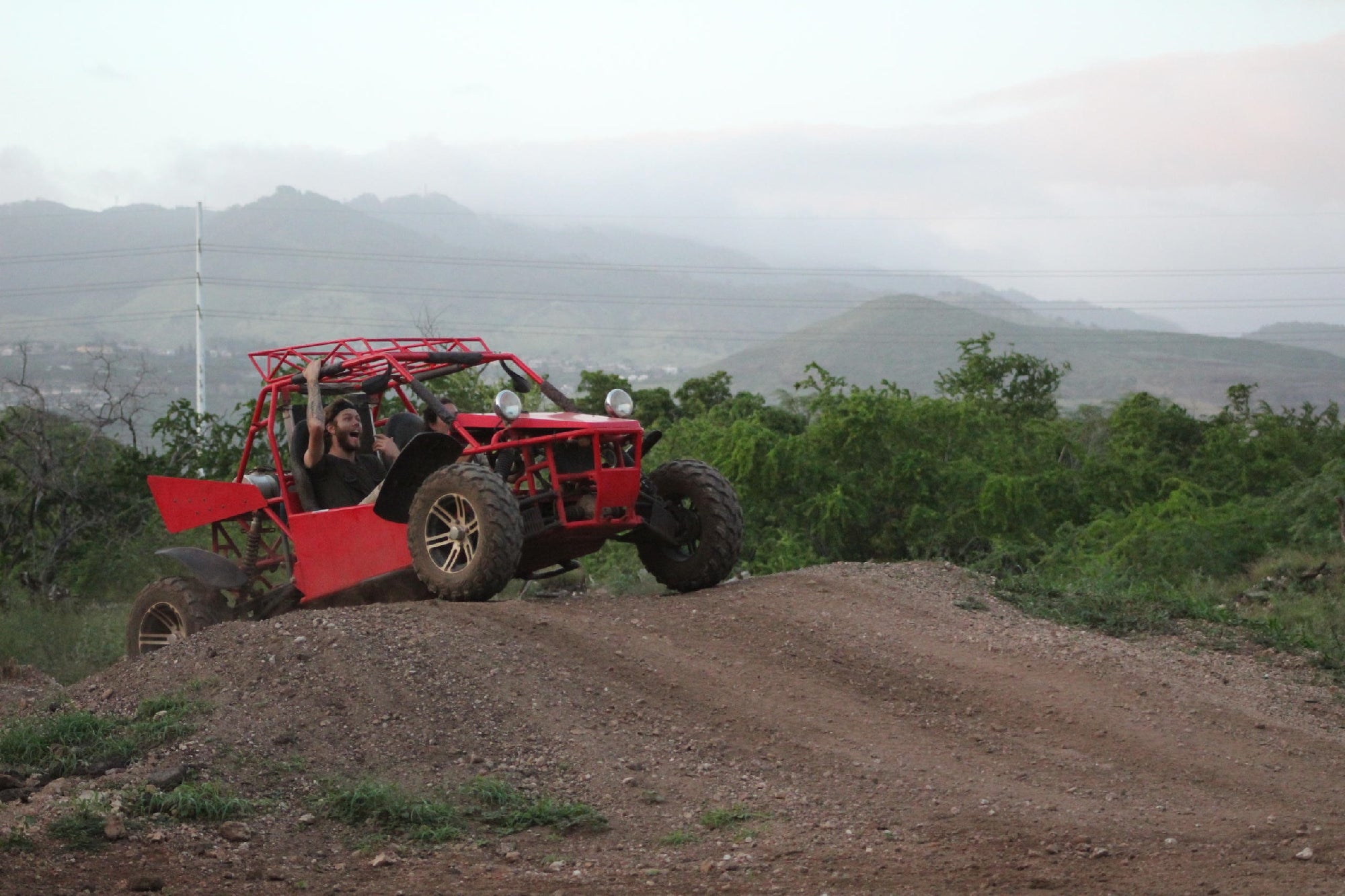 A red dune buggy with two people inside drives over a dirt mound on a rugged off-road trail with hills and greenery in the background.