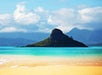A small, green island rises from turquoise water near a sandy beach with mountains and clouds in the background.