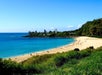 A sandy beach curves along clear blue water with people walking and relaxing; green hills and a tall building are visible in the background under a clear sky.