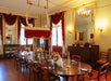 Elegant dining room with a long oval table set for a meal, surrounded by wooden chairs, floral centerpieces, red drapes, large mirrors, and a decorative chandelier.